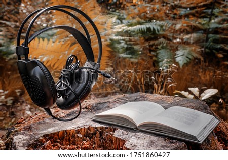 Black headphones and a white book in the forest on a stump against a background of green and orange tree leaves. Audio book reading online.