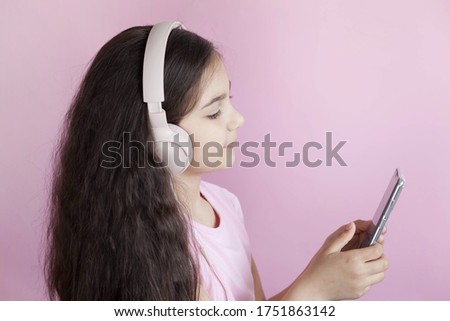 Cute child girl in headphones is using a smartphone, looking at camera and smiling on light background. Listening music. Online talking.