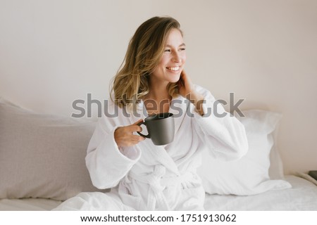 Young beautiful woman wearing white bathrobe having breakfast in bed with coffee and croissant and fresh fruits in cozy bedroom. Morning rituals. Royalty-Free Stock Photo #1751913062