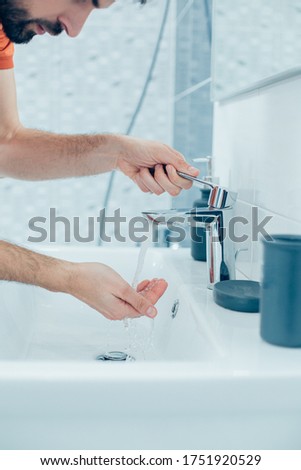 Close up photo of a young man leaning to the bathroom basin and putting his hands under the clean running water