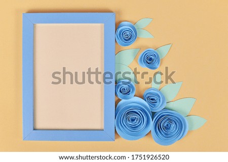 Flower flat lay with empty picture frame surrounded by romantic blue paper craft roses with leaves on yellow background