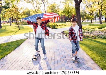Two cute boys brothers riding skateboards in a public park on a sunny afternoon. U.S. Independence Day. The older brother keeping the waving flag of America in the hands. Showing big fingers up.