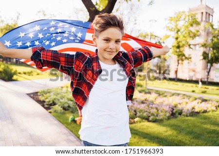 U.S. Independence Day. A young guy American or caucasian, 8 years old riding at the speed on a skateboard. Holding the fluttering flag of USA America. Celebrating July 4th. Adorable little kid.