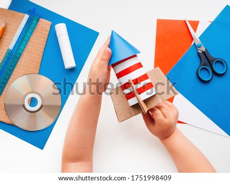 Child makes colorful rocket out of colored paper. Also he needs carton, scissors, glue, CD-disk, pencil and ruler. Red, white and blue handmade spaceship. Making toys from improvised materials.