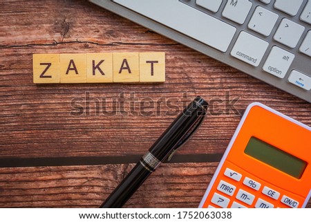 Conceptual zakat (islamic tax) with fountain pen, keyboard and calculator. Obligation to aid the poors. ZAKAT text on wooden table. Islamic zakat concept. Selective focus.