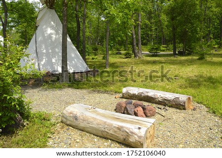 Two wooden seats made of bars next to a bonfire of stones on the background of native American wigwam in forest Royalty-Free Stock Photo #1752106040