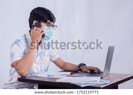 Young asian man answer calling the phone during work from home and wearing face mask to protect his life from corona virus