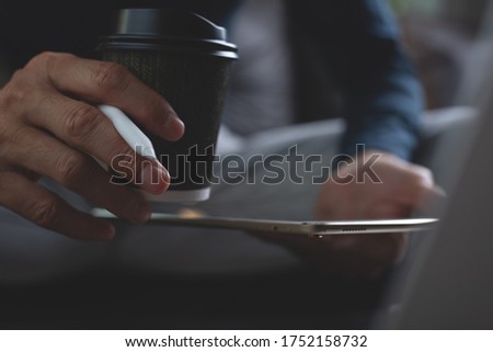 Close up of casual man, hipster holding take home cup of coffee, online working on digital laptop computer. Graphic designer with stylus pen working on digital tablet and drinking coffee