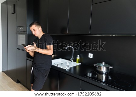 Smiling Caucasian young man use cellphone gadget texting messaging standing in kitchen
