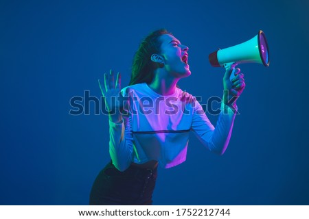 Shouting with speaker. Caucasian young woman's portrait isolated on blue studio background in neon light. Beautiful female model. Concept of human emotions, facial expression, sales, ad, youth culture