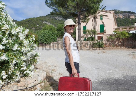 Wanderlust concept of  young woman walking through the field in spring summer wearing a hat and glasses holding a vintage red suitcase in her hand, her face expresses happiness and she is on vacation.