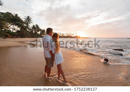 Young happy couple on seashore, looking beautiful golden sunset during vacation on tropical island. Palm trees on background, waves touch their feets