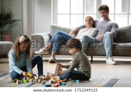 Little preschooler children sit on warm floor in living room play construct with colorful building blocks bricks together, young parents rest on sofa in background, Caucasian family relax at home