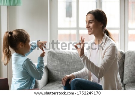 Smiling young Caucasian mom and little daughter make hand gesture learn speak sign language at home, happy mother or nanny practice nonverbal talk with small disabled girl child, disability concept