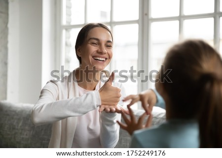 Smiling young Caucasian mom or nanny make hand gesture practice nonverbal talk with little girl child, positive female teacher or tutor learn sign language with small disabled kid, disability concept