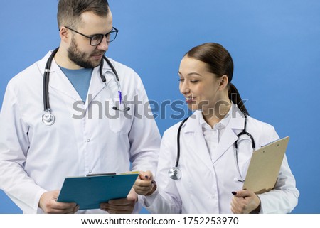 Two positive male and female doctors in white lab coat and stethoscope analyzing data on clipboard on blue background. Healthcare, hospital, medical clinic workers, insurance, medicine career concept