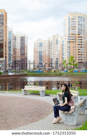 Relaxed young woman in a green T-shirt and with a backpack sitting on a wooden bench, drinking coffee and browsing on her laptop