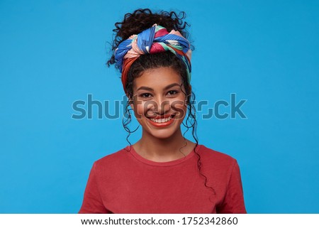 Happy young attractive dark haired curly woman with multi-colored headband showing her white perfect teeth while smiling cheerfully, isolated over blue background