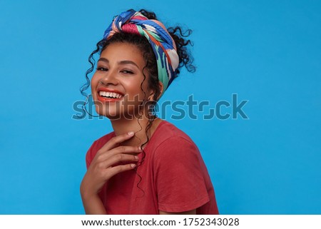Studio photo of young attractive joyful brunette curly lady keeping her hand raised and looking gladly at camera with broad smile, isolated over blue background