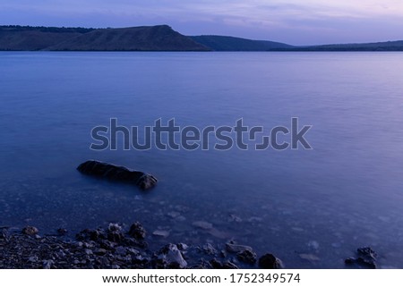 landscape evening long exposure river shore line with soft focus stone in shallow water foreground and hill land background horizon space in twilight lighting after sunset 