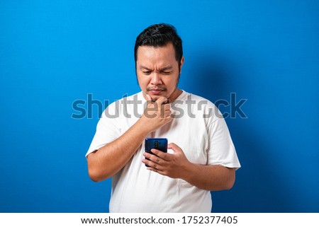 Fat Asian man in white t-shirt, thinking something while chat messaging on his smart phone, half body portrait over blue background