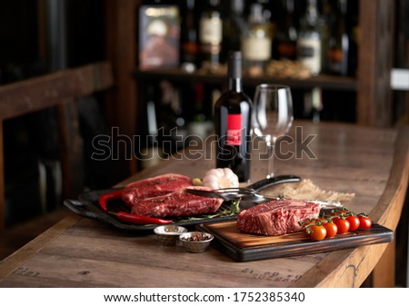   Two juicy, fresh striploin steaks in a grill pan and ribeye steak on a cutting board on a countertop and barrel boards with tomatoes on a branch, peppercorns, red chili peppers, a meat fork 