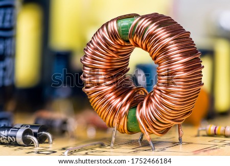 Induction coil with copper wire winding soldered on printed circuit board. Toroidal inductor with magnetic ferrite core. Electronic components on switch-mode power supply unit detail. Selective focus. Royalty-Free Stock Photo #1752466184
