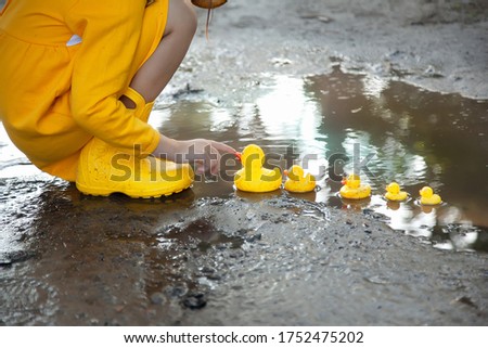 A girl in a yellow dress and yellow rubber boots lets a family of yellow ducks in a puddle. Bright picture of summer holidays. Summer rain. Spring

