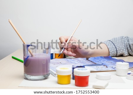 Female hand holds a brush and draws picture at table in middle of jars with watercolor paints and next to it is a glass for rinsing and cleaning the brush from color on a white background in studio.