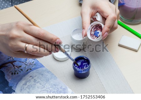 A close-up of female hands over a painted picture holding a brush and a cover for watercolors on which the colors are mixed next to the eraser and a simple pencil. A hobby of art.