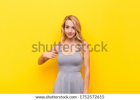 young blonde woman looking proud, confident and happy, smiling and pointing to self or making number one sign against orange wall