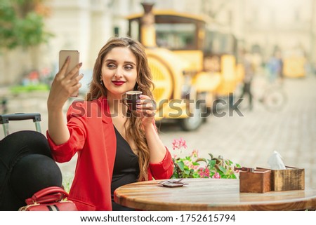 Beautiful brunette woman taking pictures of herself on a cellphone holding cup of coffee at cafe outdoors.