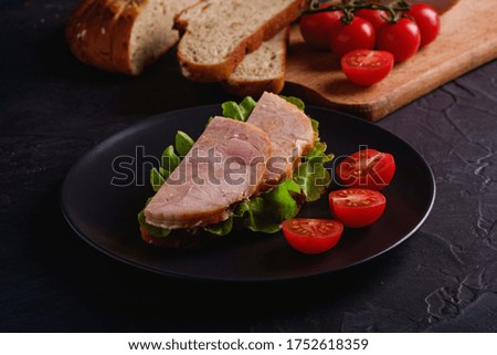 Sandwich with turkey ham meat, green salad and fresh cherry tomatoes slices on black plate near to ingredients on cutting board, dark textured background, angle view