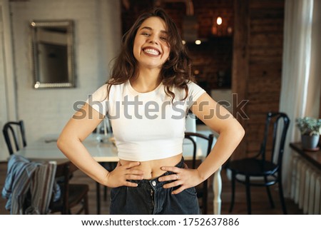 Leisure, relaxation, home and domesticity. Positive cheerful teenage girl wearing crop top smiling broadly at camera, having fun indoors, spending day at home, keeping hands on her waist, laughing