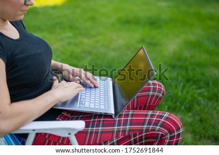 woman sits in a park and types on the computer while shopping online. The girl maintains a social distance and works outdoors on a laptop. The student is studying remotely.