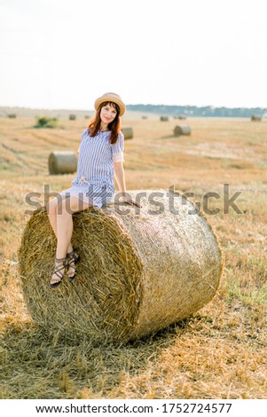 Countryside landscape of farm wheat field at summer sunset. Pretty young happy Caucasian woman in straw hat and blue striped dress sitting on hay bale and enjoying warm evening and vacations
