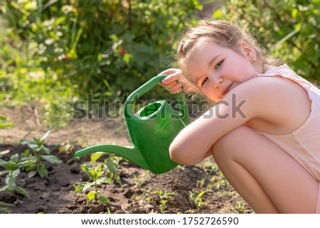Lovely smiling little girl outdoor in garden takes care of plants by watering it by green watering can. Activities with children outdoors. Summer, spring, gardening concept. Copy space.