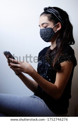 Soft focus of a beautiful brunette teen girl in a black shirt using a mobile phone at home with a cloth mask over light gray wall background.