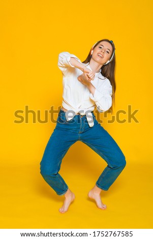 Joyous american woman in casual clothing dancing and listening to music with pleasure via white earphones isolated over yellow background