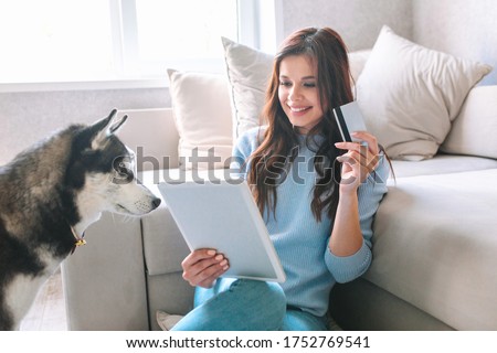 Young woman holding credit card and using tablet with her dog at home. Online shopping, e-commerce, internet banking,
