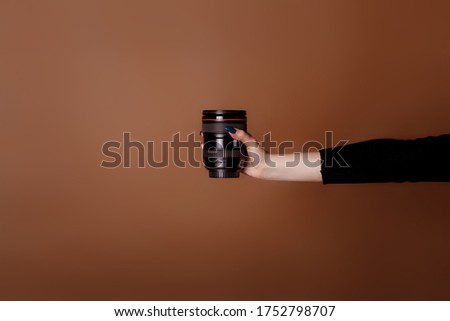 Female hand holds a lens on a brown background. World photography day. Studio photography. Space for text. Copy space