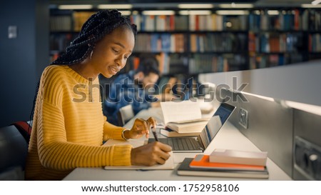 University Library: Gifted Beautiful Black Girl Sitting at the Desk, Uses Laptop, Writes Notes for the Paper, Essay, Study for Class Assignment. Diverse Group of Students Learning, Studying for Exams. Royalty-Free Stock Photo #1752958106