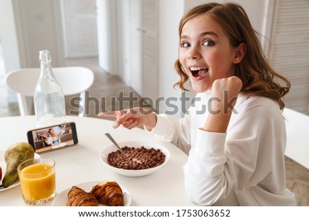 Photo of joyful girl taking video call and pointing finger at cellphone while having breakfast in kitchen