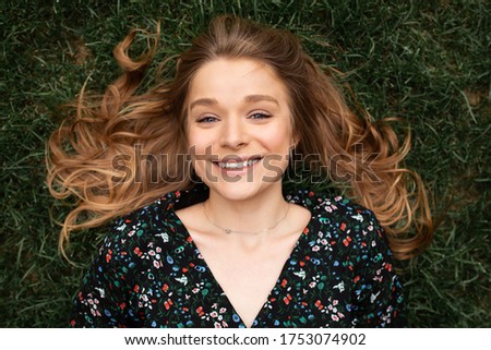 Young beautiful cheerful woman lying alone on green grass and smile on camera. Girl looks straight and poses. Her blonde har on green grass. Picture concept