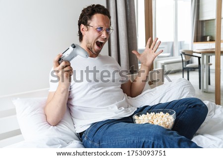 Handsome cheerful young man playing on playstation while laying on bed at home, eating popcorn, celebrating