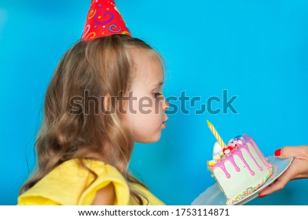 Young cute celebrating girl with curly long hair and birthday cap blowing candle while her mom's hand holding a piece of cake on blue background. Copy space