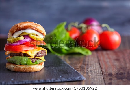A big, fat, juicy double cheeseburger made with two 100% beef patties, slices of melted cheese, onions, pickles, lettuce, tomatoes, and mayo on a fresh sesame seed bun over a rustic dark background. 