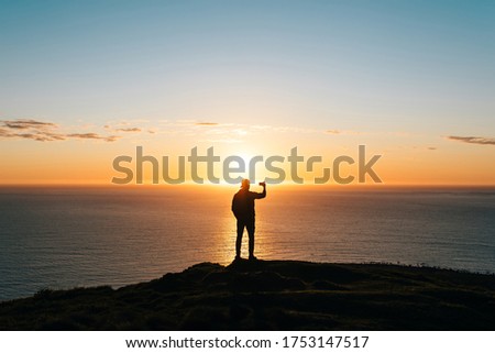Silhouette of a young man watching sunset at the seaside and taking pictures with phone camera. Travel concept. 