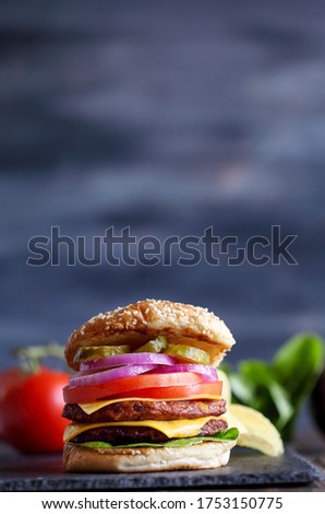 Vegetarian black bean cheeseburger made with two meat substitute patties, slices of melted cheese, onions, pickles, lettuce, and tomato on a fresh sesame seed bun over a rustic dark background. 
