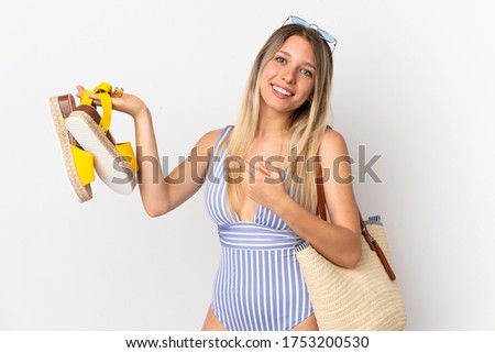 Young blonde woman holding summer sandals isolated on white background and pointing it